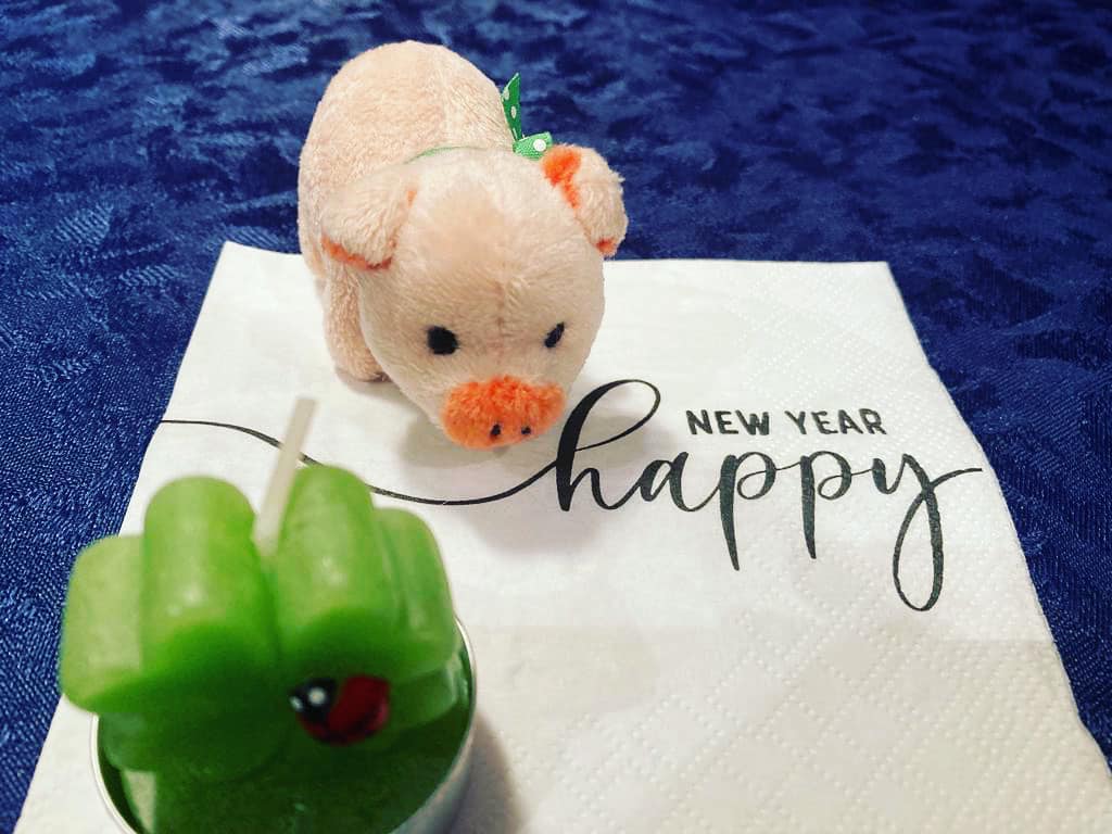 Auf einer Serviette mit der Aufschrift "Happy New Year" stehen ein kleines weißes Glücksschwein und eine Kerze in Form eines vierblättrigen Kleeblatts